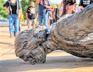 A crowd stands near a fallen statue of a wavy-haired man, his nose resting on the pavement.