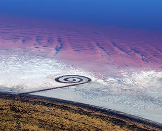 An aerial view of a spiral-shaped land art sculpture made out of mud, salt crystals, and balsalt rocks. An aerial view of a spiral-shaped land art sculpture made out of mud, salt crystals, and balsalt rocks.