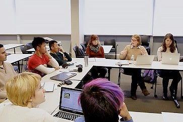 Seven students and an instructor sit around a U-shaped table. The instructor is speaking, and several students look in her direction. 