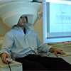 Photo of a person sitting in a chair with a large cylindrical apparatus around their head, looking at a computer screen and holding a response device.