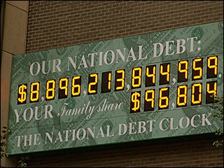 A photograph of a billboard sized electronic clock on a brick wall that displays the national debt of the United States.