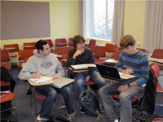 Three students sitting at desks, reading to each other from textbooks.