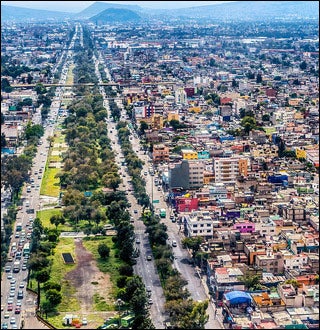 An aerial photograph showing buildings crowded together on the right side with mountains in the background.  A long thin strip of green runs parallel to the roads.