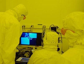 People in cleanroom suits view magnified wafer pattern on screen.