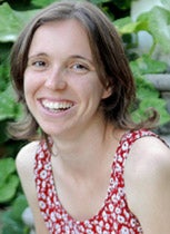 A young woman smiles brightly at the camera. She is sitting in front of some green plants and is wearing a bright red and white shirt.