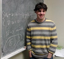 A young man with black hair is standing beside a chalkboard with equations on it. He is wearing a sweater with grey and yellow horizontal stripes. His hands are in his pockets.