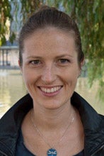 A woman in a black coat with a blue necklace stands in front of a pond.