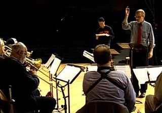 Photo of part of a large jazz ensemble on stage - saxophones and trombones - with conductor and composer out front.