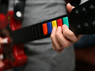 A person playing a Guitar Hero controller that looks like a red electric guitar with multi-colored buttons instead of strings.