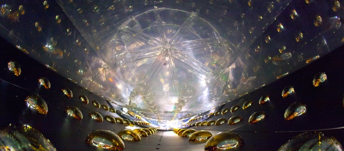 An array of bright yellow hemispheres line the outside of a irregularly shaped dark metal chamber, with a lit transparent element at the bottom.