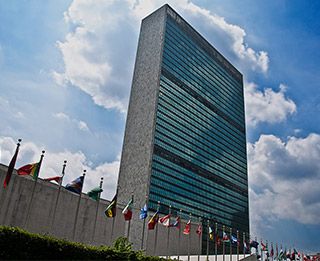 A tall glass and concrete structure fronted by the flags of various nations stands against a blue sky.