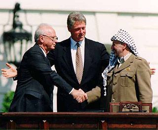 Two gray-haired gentleman, one wearing a suit, the other wearing a brown jacket and a long scarf, shake hands while a younger man stands between them.