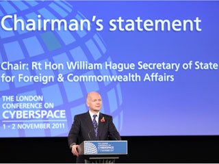 A slim, formally dressed balding man stands at a podium in front of a huge projection screen.