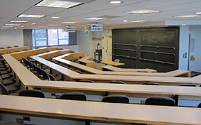 Five rows of lecture tables with grey chairs; black sliding boards at the front of the room; a window on the left wall.