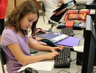 A young girl in a lilac shirt sits at a computer working on homework.