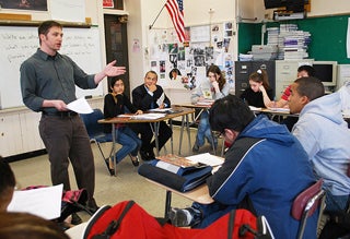 A teacher stands in a classroom with students seated and taking notes. The governor of Massachusetts sits with the students.