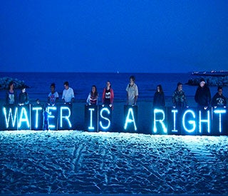 Protesters stand on a beach at nightfall with a neon blue sign that reads “Water is a right”.