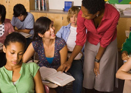 Students sit at desks in rows. A teacher leans over to help a student with an assignment.