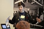 A photo of a woman holding a piece of paper, talking at the front of a room.