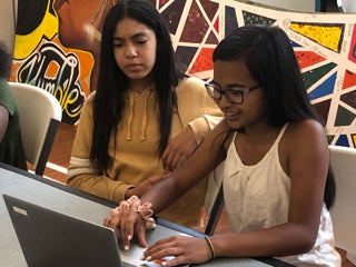 Two girls sitting in front of a laptop, with one typing on laptop and the other girl looking at the laptop screen