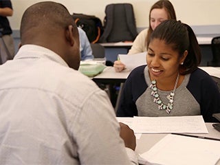 Photo of part of a classroom where 2 students are talking and more students behind them.