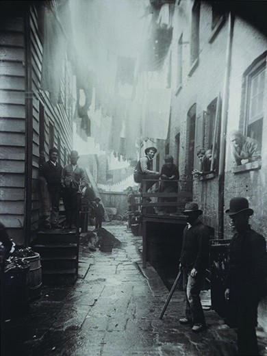 A photograph shows an alley between two tenements. Men, women, and children stand on either side of the alley, in the stoops, and in the windows